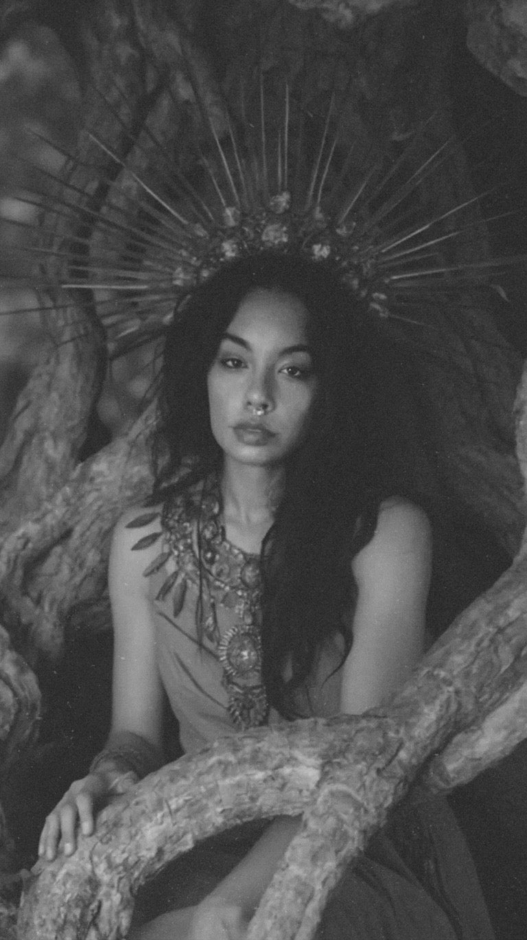 Woman in ornate dress with large decorative headpiece sitting among stone and foliage
